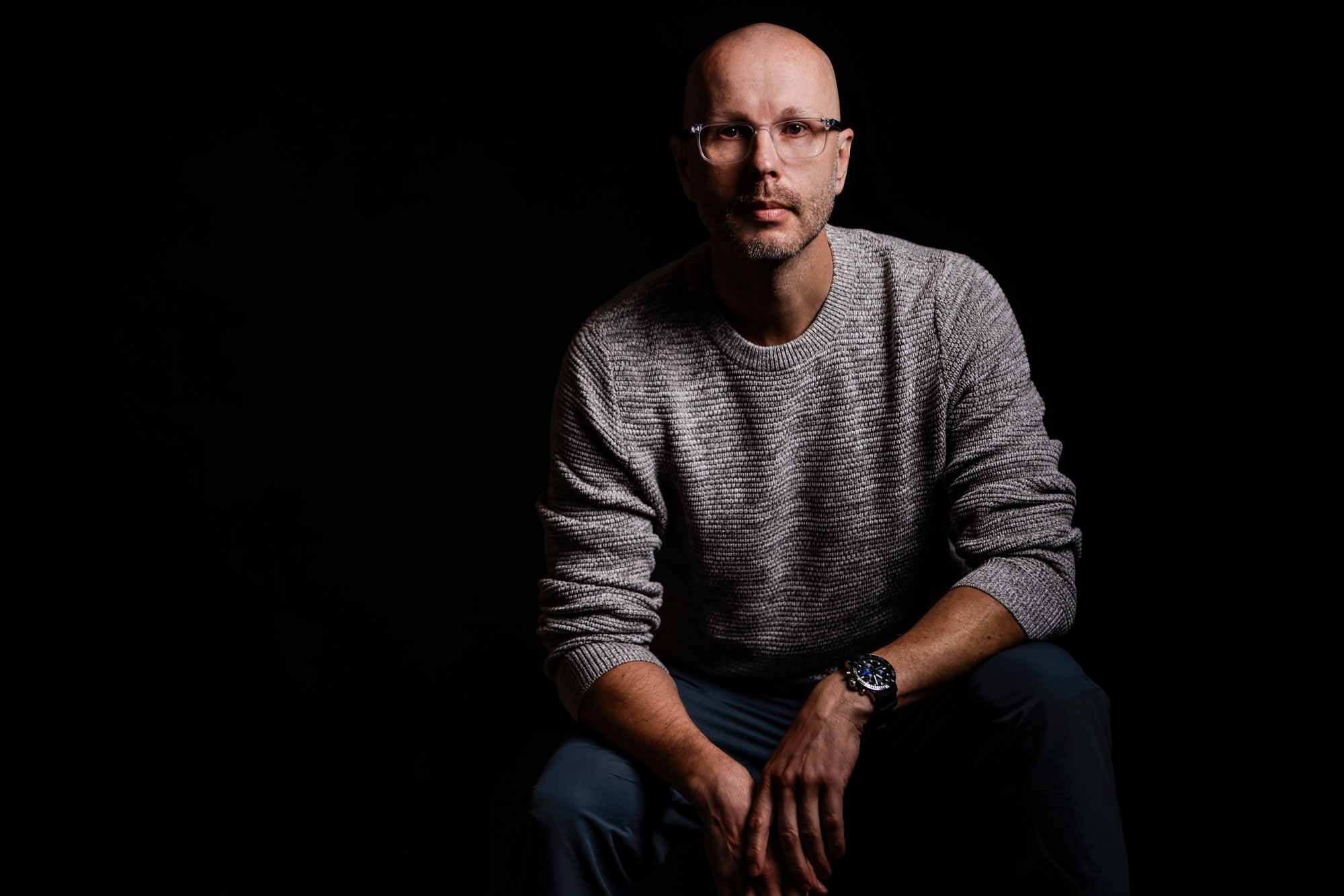 Foto von Axel Hillebrandt - Porträt mit Brille im grauen Pullover vor schwarzem Hintergrund, sitzend mit nach vorne gestützten Armen.