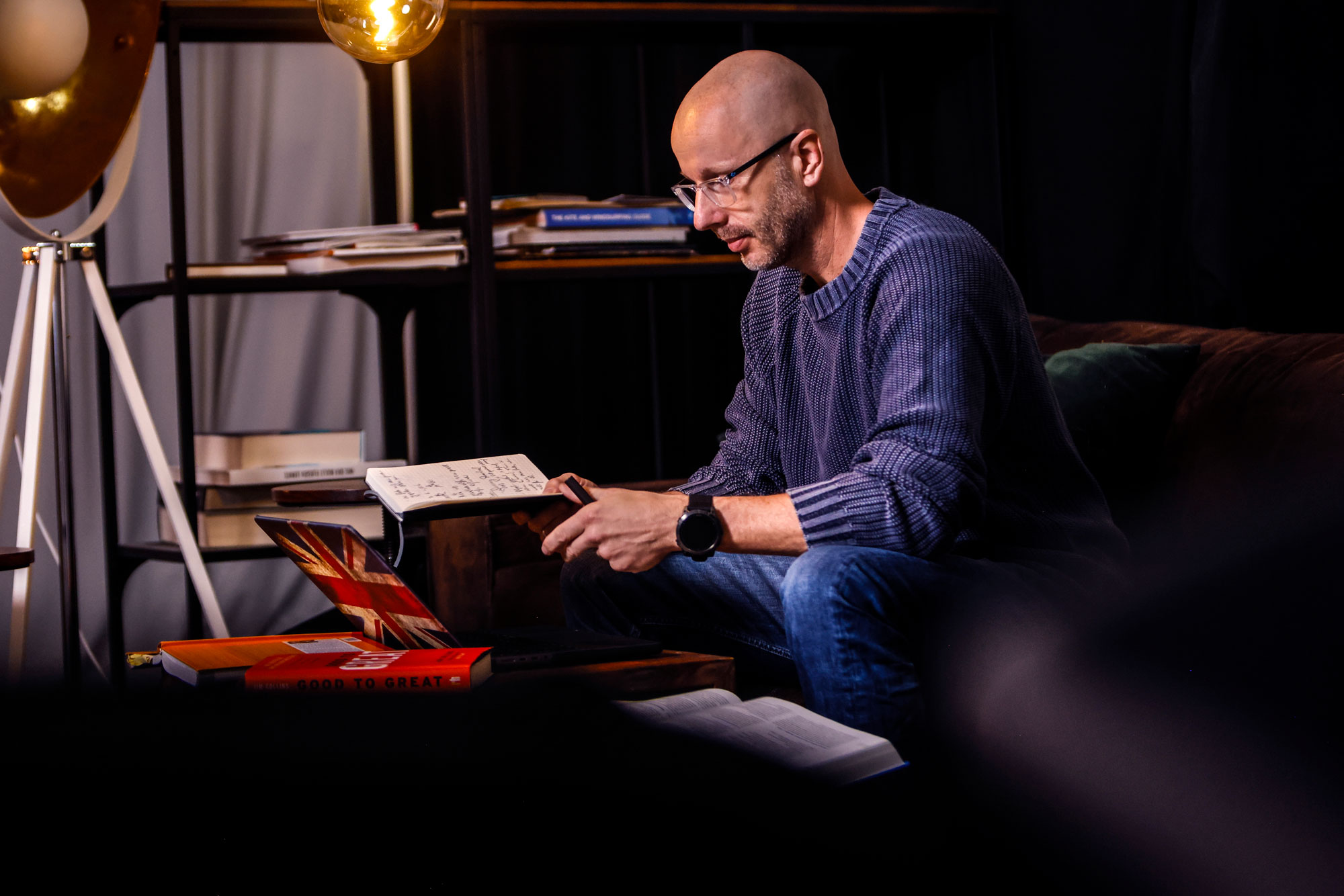 Foto von Axel Hillebrandt auf einem Sofa in warm ausgeleuchtetem Raum, der in einem Notizbuch schreibt, umgeben von Büchern und einem Laptop.